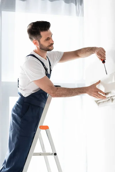 Handsome repairman standing on stepladder and fixing air conditioner with screwdriver — Stock Photo