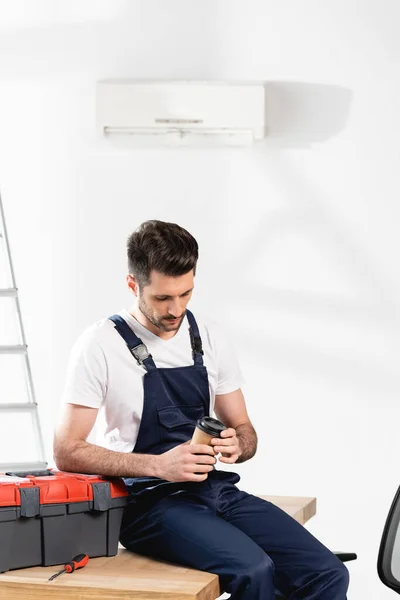 Junger Reparateur mit Coffee to go, während er am Schreibtisch in der Nähe des Werkzeugkastens unter Klimaanlage an der Wand sitzt — Stockfoto