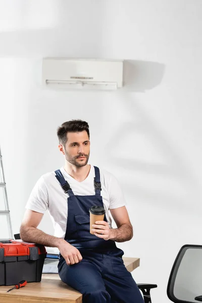 Reparador guapo sosteniendo café para ir sentado en el escritorio cerca de la caja de herramientas bajo el aire acondicionado en la pared - foto de stock
