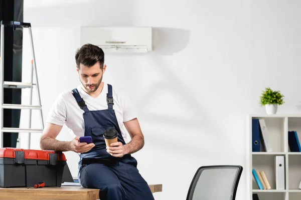 Joven trabajador sentado en el escritorio, el uso de teléfono inteligente y la celebración de café para ir cerca de aire acondicionado - foto de stock