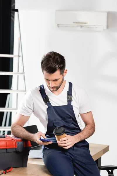 Reparador sentado na mesa, usando smartphone e segurando café para ir perto de ar condicionado na parede — Fotografia de Stock