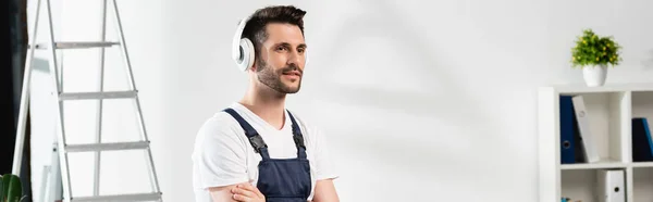 Horizontal image of repairman in wireless headphones standing with crossed arms near stepladder and air conditioner on wall — Stock Photo