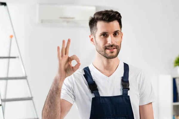 Lächelnder Reparateur zeigt Okay-Geste, während er neben Leiter und Klimaanlage an der Wand steht — Stockfoto