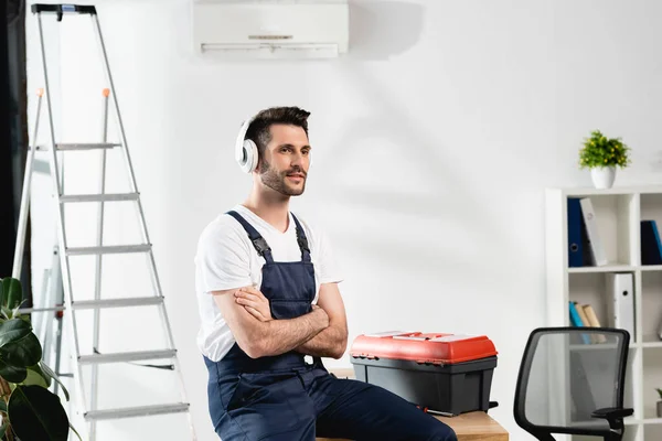 Trabajador en auriculares inalámbricos sentado en el escritorio con brazos cruzados cerca de la caja de herramientas y aire acondicionado en la pared - foto de stock