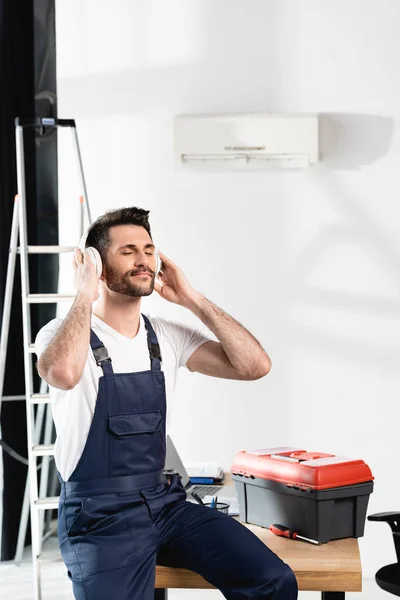 Reparateur mit geschlossenen Augen sitzt auf Schreibtisch in drahtlosen Kopfhörern in der Nähe von Werkzeugkiste und Klimaanlage an der Wand — Stockfoto