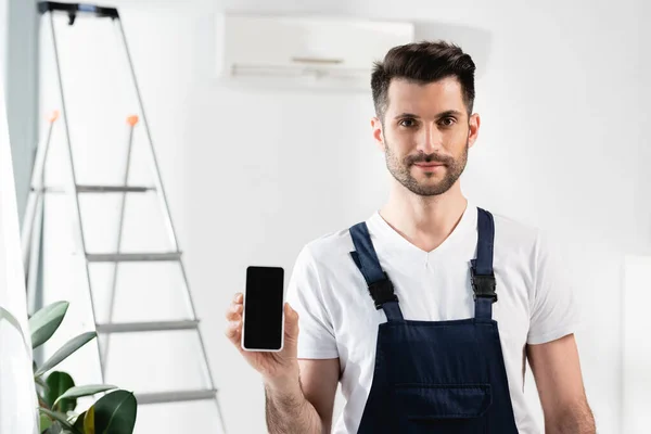 Reparador guapo mostrando teléfono inteligente con pantalla en blanco cerca de escalera y aire acondicionado en la pared - foto de stock