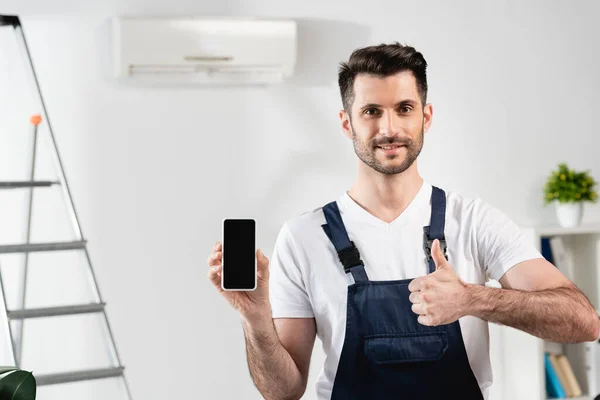 Reparador sonriente sosteniendo teléfono inteligente con pantalla en blanco y mostrando el pulgar hacia arriba - foto de stock