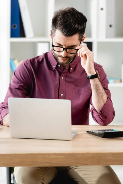 Homem de negócios sério tocando óculos enquanto trabalhava no laptop no escritório — Fotografia de Stock