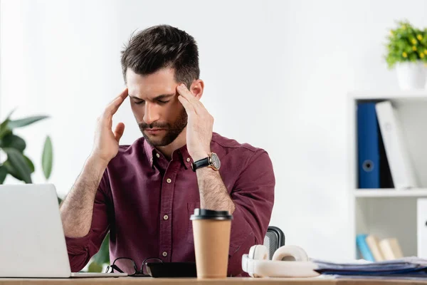 Erschöpfter Geschäftsmann mit geschlossenen Augen, der den Kopf berührt, während er neben Laptop und Coffee to go sitzt — Stockfoto