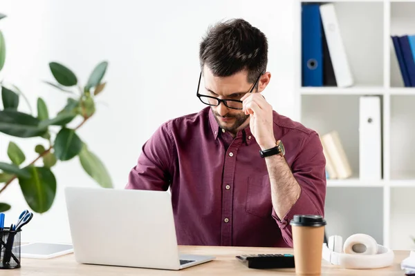 Серйозний бізнесмен торкається окулярів, сидячи на робочому місці біля ноутбука — стокове фото