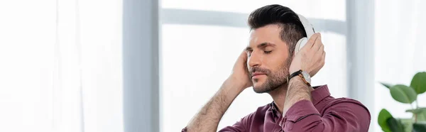 Horizontal image of dreamy businessman listening music in wireless headphones in office — Stock Photo