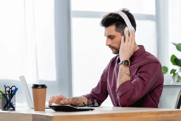 Homem de negócios concentrado em fones de ouvido sem fio digitando no laptop perto de café para ir — Fotografia de Stock