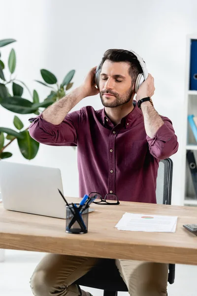 Nachdenklicher Geschäftsmann mit geschlossenen Augen, der am Arbeitsplatz drahtlose Kopfhörer anfasst — Stockfoto
