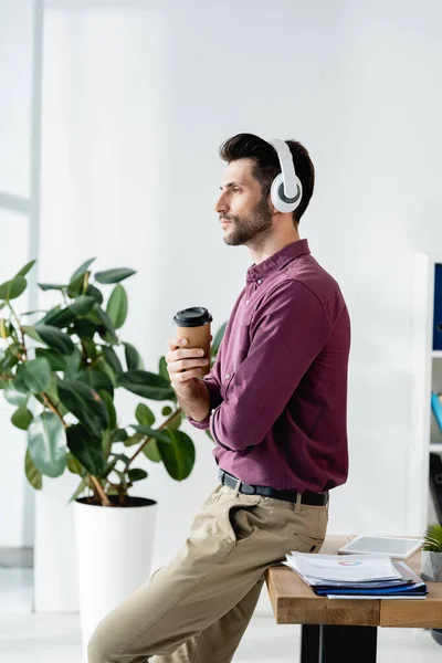 Vista laterale di uomo d'affari premuroso in cuffie wireless che tengono il caffè per andare mentre seduto sulla scrivania vicino alla pianta in vaso — Foto stock