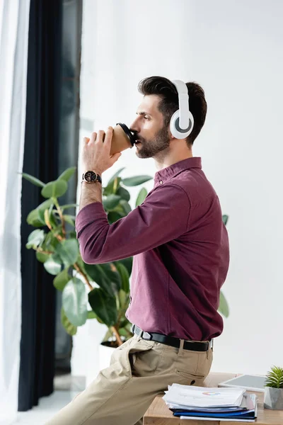 Vista lateral del hombre de negocios bebiendo café para ir mientras está sentado en el escritorio - foto de stock