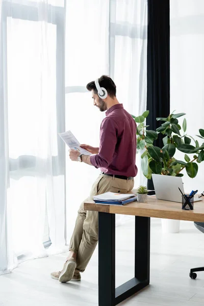 Businessman in wireless headphones looking at document while sitting on desk near window — Stock Photo