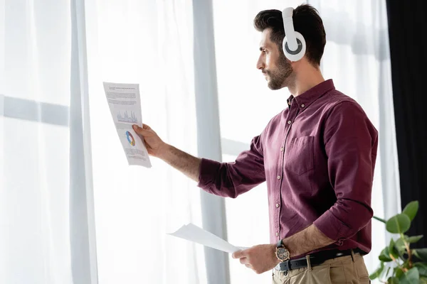 Hombre de negocios en auriculares inalámbricos mirando papel con análisis de negocios cerca de ventana - foto de stock