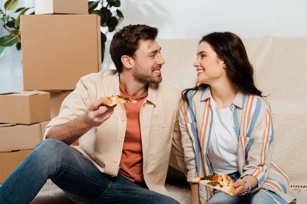 Пара улыбается друг другу, держа кусочки пиццы возле картонных коробок в гостиной — стоковое фото