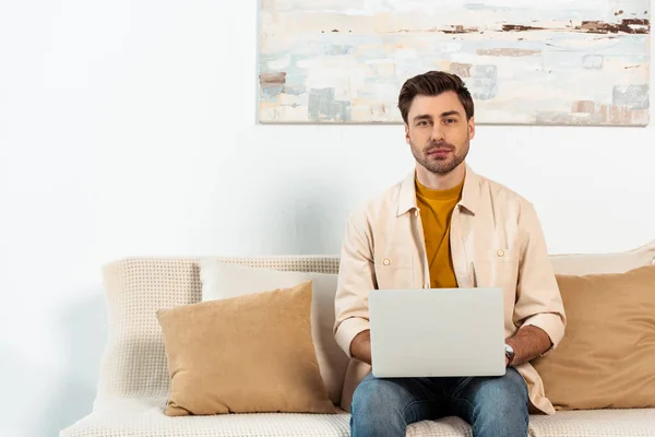 Schöne Freiberuflerin blickt in die Kamera, während sie Laptop auf der Couch hält — Stockfoto