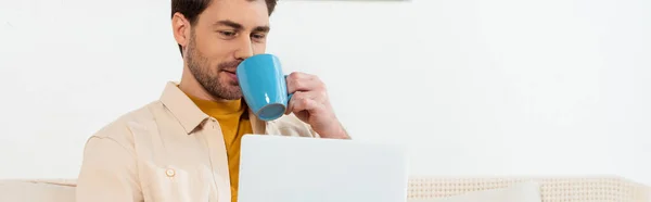 Foto panorámica del hombre guapo bebiendo café y utilizando el ordenador portátil en casa - foto de stock