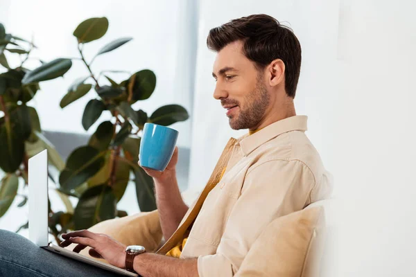 Concentration sélective du bel homme tenant une tasse de café et utilisant un ordinateur portable à la maison — Photo de stock