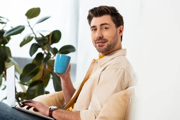 Вид сбоку на красивого фрилансера, держащего чашку кофе и использующего ноутбук в гостиной — стоковое фото