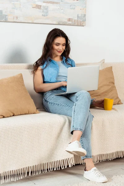 Mulher bonita usando laptop e segurando xícara de café no sofá — Fotografia de Stock