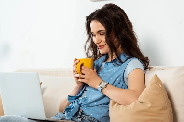 Sonriente freelancer sosteniendo la taza y mirando el portátil en el sofá - foto de stock