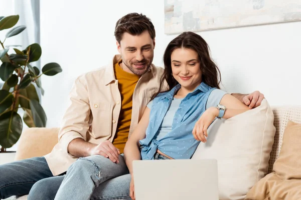 Selective focus of handsome man sitting near smiling girlfriend and laptop on couch — Stock Photo