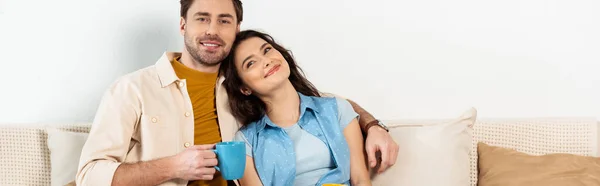 Панорамний знімок красивого чоловіка тримає чашку кави і обіймає усміхнену дівчину на дивані — стокове фото