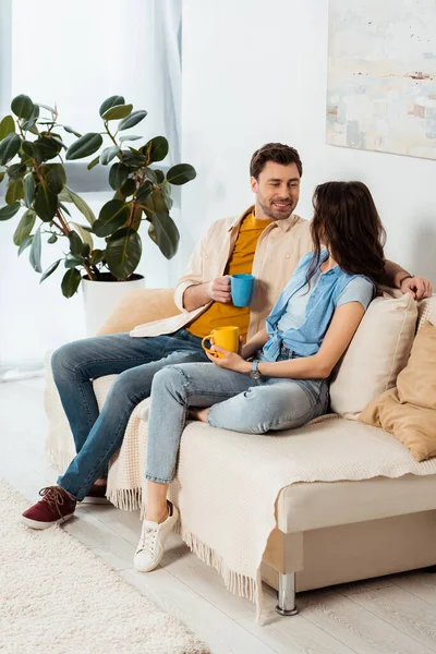 Beau homme souriant à sa copine tout en tenant une tasse de café sur le canapé — Photo de stock