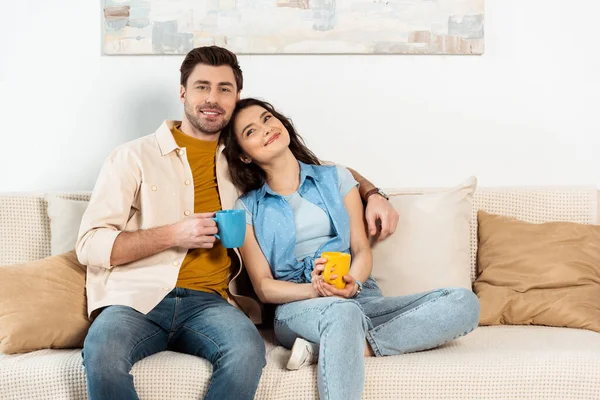 Handsome man smiling at camera while embracing girlfriend and holding cup of coffee on sofa — Stock Photo