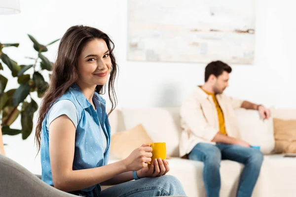 Enfoque selectivo de la mujer sosteniendo la taza de café y sonriendo a la cámara cerca de novio en el sofá - foto de stock