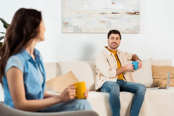 Enfoque selectivo del hombre sosteniendo la taza de café cerca de la computadora portátil y sonriendo a la novia en la sala de estar - foto de stock