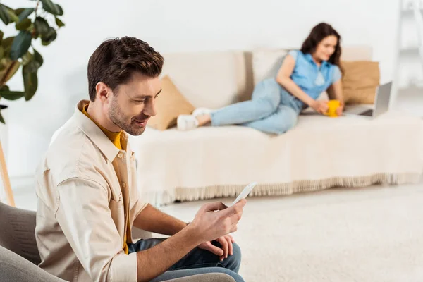 Selective focus of smiling man using smartphone while girlfriend with cup and laptop lying on couch — Stock Photo
