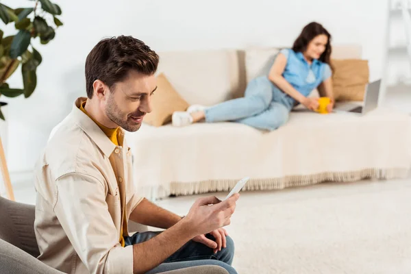 Focus selettivo di uomo sorridente utilizzando smartphone mentre fidanzata con tazza e laptop sdraiato sul divano — Foto stock