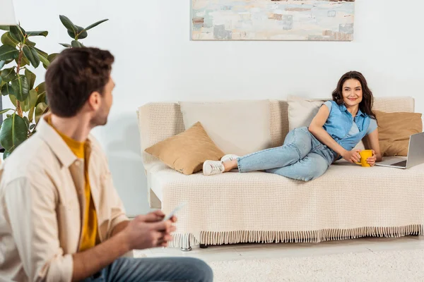 Selective focus of woman holding cup near laptop on couch and smiling at boyfriend in living room — Stock Photo