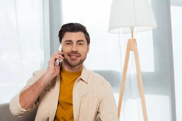 Smiling man looking at camera while talking on smartphone at home — Stock Photo