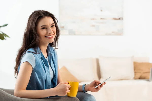 Ragazza attraente sorridente alla fotocamera durante l'utilizzo di smartphone e tenendo la tazza di caffè in soggiorno — Foto stock