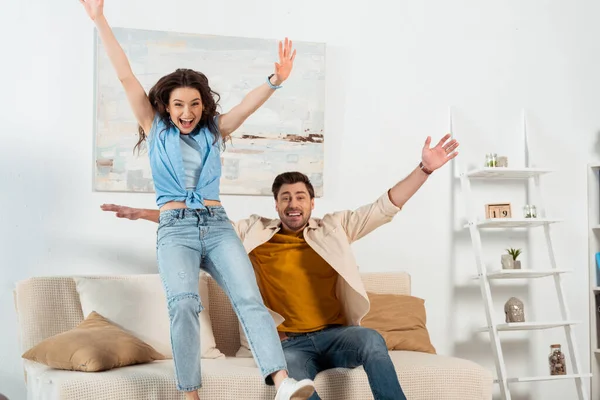 Fröhliches Paar mit erhobenen Händen blickt im Wohnzimmer in die Kamera — Stockfoto