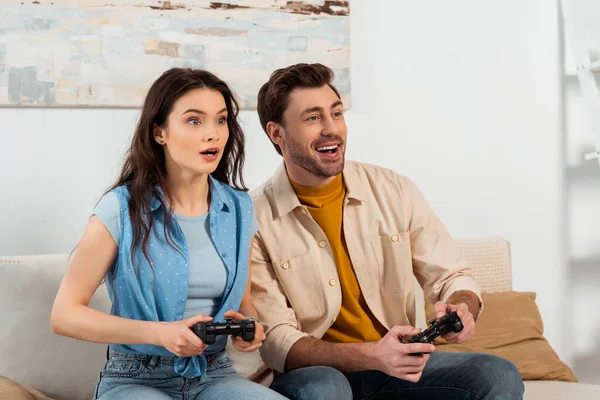 KYIV, UCRANIA - 4 de junio de 2020: Una mujer conmocionada jugando un videojuego cerca de su novio sonriente en casa - foto de stock