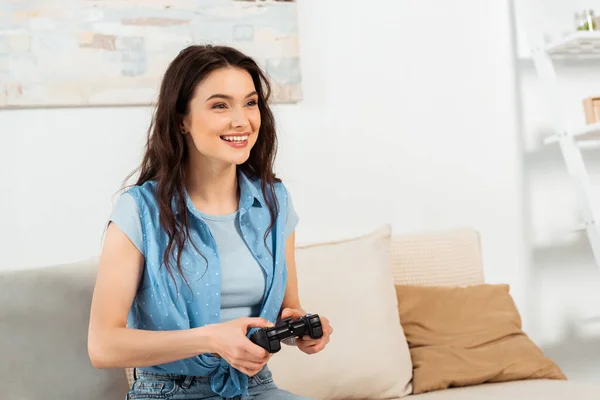 KYIV, UCRANIA - 4 de junio de 2020: Atractiva chica sonriente sosteniendo el joystick mientras juega un videojuego en casa - foto de stock
