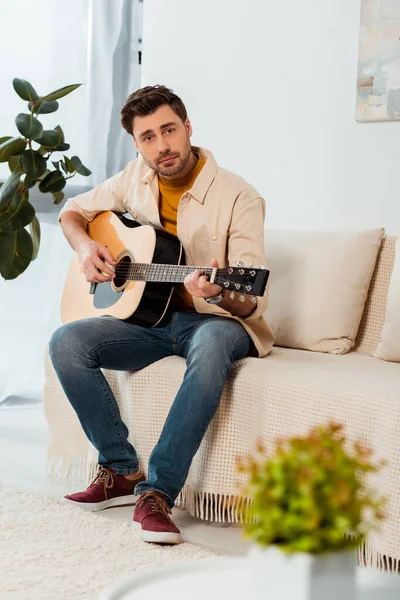 Вибірковий фокус людини дивиться на камеру, граючи на акустичній гітарі вдома — стокове фото