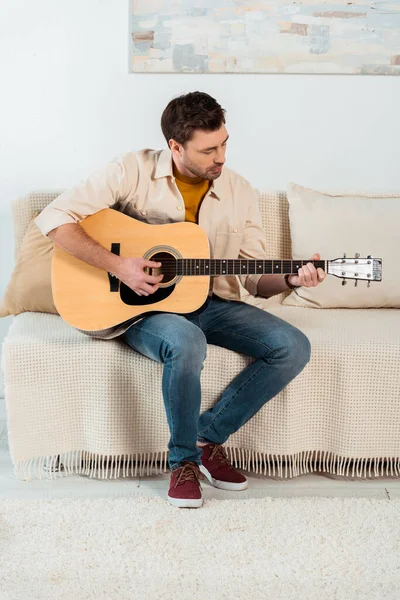 Young man performing on acoustic guitar on sofa in living room — Stock Photo