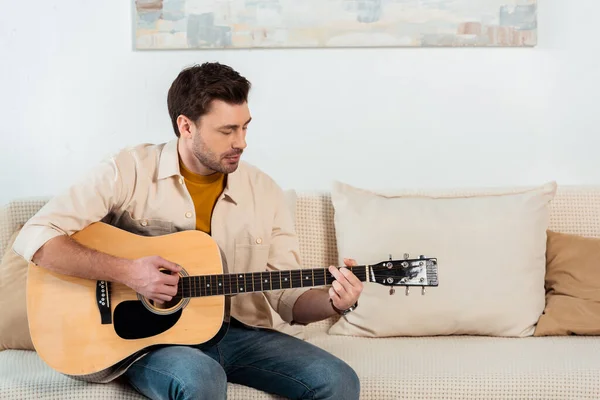 Hombre guapo tocando la guitarra acústica en el sofá en la sala de estar - foto de stock