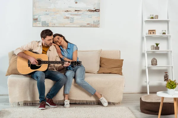 Красивый мужчина играет на акустической гитаре рядом с улыбающейся женщиной на диване дома — стоковое фото
