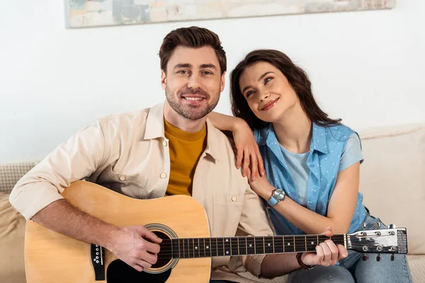 Hombre guapo sonriendo a la cámara mientras toca la guitarra acústica cerca de su novia - foto de stock