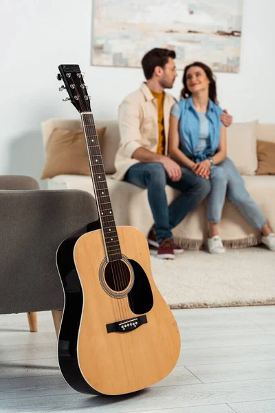 Concentration sélective de la guitare acoustique sur le sol près du fauteuil et l'homme embrassant petite amie à la maison — Photo de stock