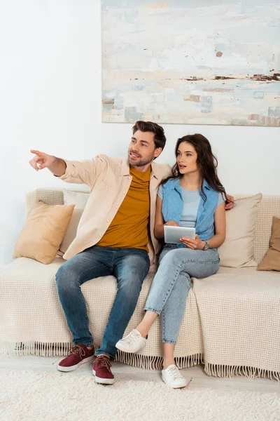 Homme souriant pointant du doigt près de la petite amie en utilisant une tablette numérique à la maison — Photo de stock