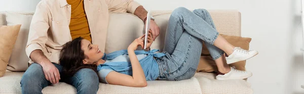Панорамний урожай усміхненої дівчини за допомогою цифрового планшета, лежачи біля чоловіка на дивані вдома — стокове фото